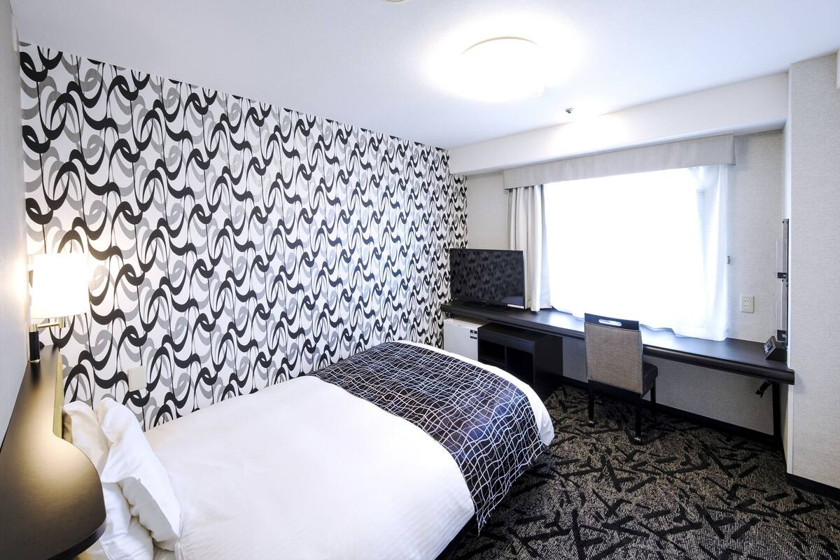 Une chambre d’hôtel avec un lit, un bureau avec une télévision, une lampe et une fenêtre avec des rideaux.