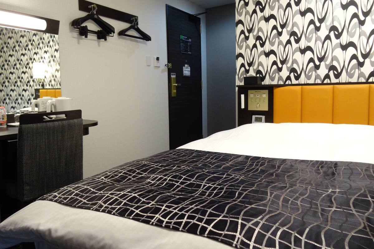 A hotel room with a bed, a desk with a chair, a wardrobe, and a door with a digital clock.