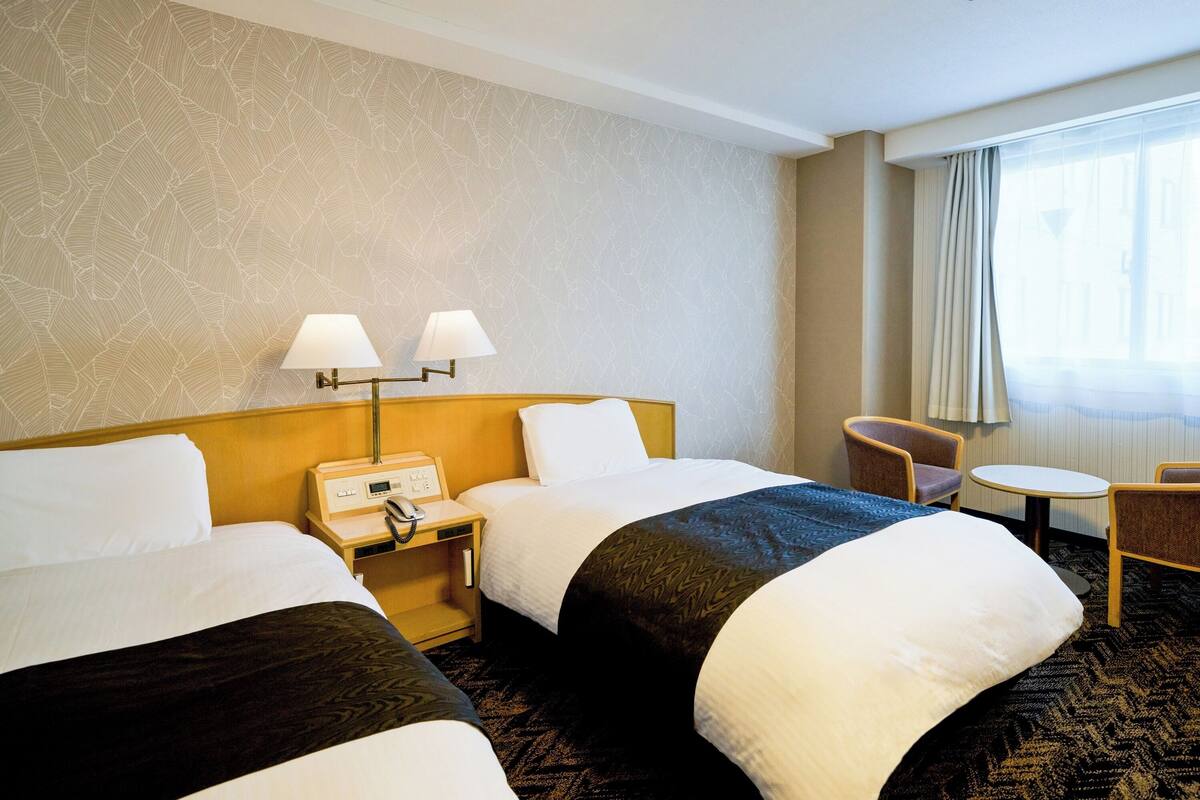A hotel room with two beds, a small table, a chair, and a nightstand with a phone.