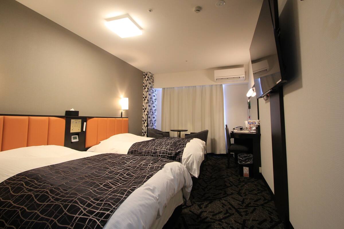 A hotel room with two beds, a desk, a chair, a TV, and an air conditioning unit.