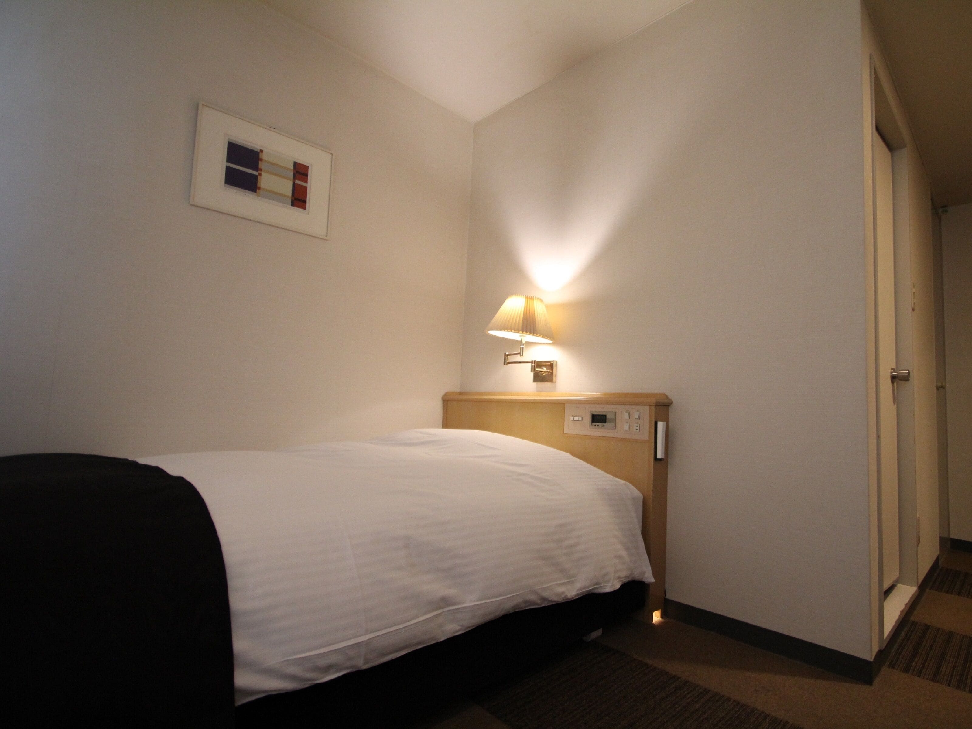 A hotel room with a bed, a bedside lamp, a control panel, and a framed picture on the wall.