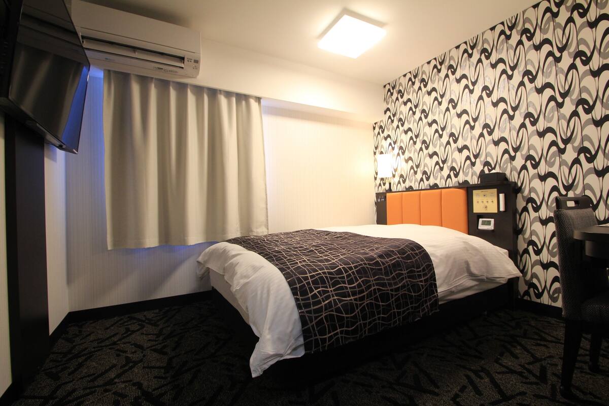A hotel room with a bed, air conditioning unit, curtains, patterned wallpaper, and a desk.