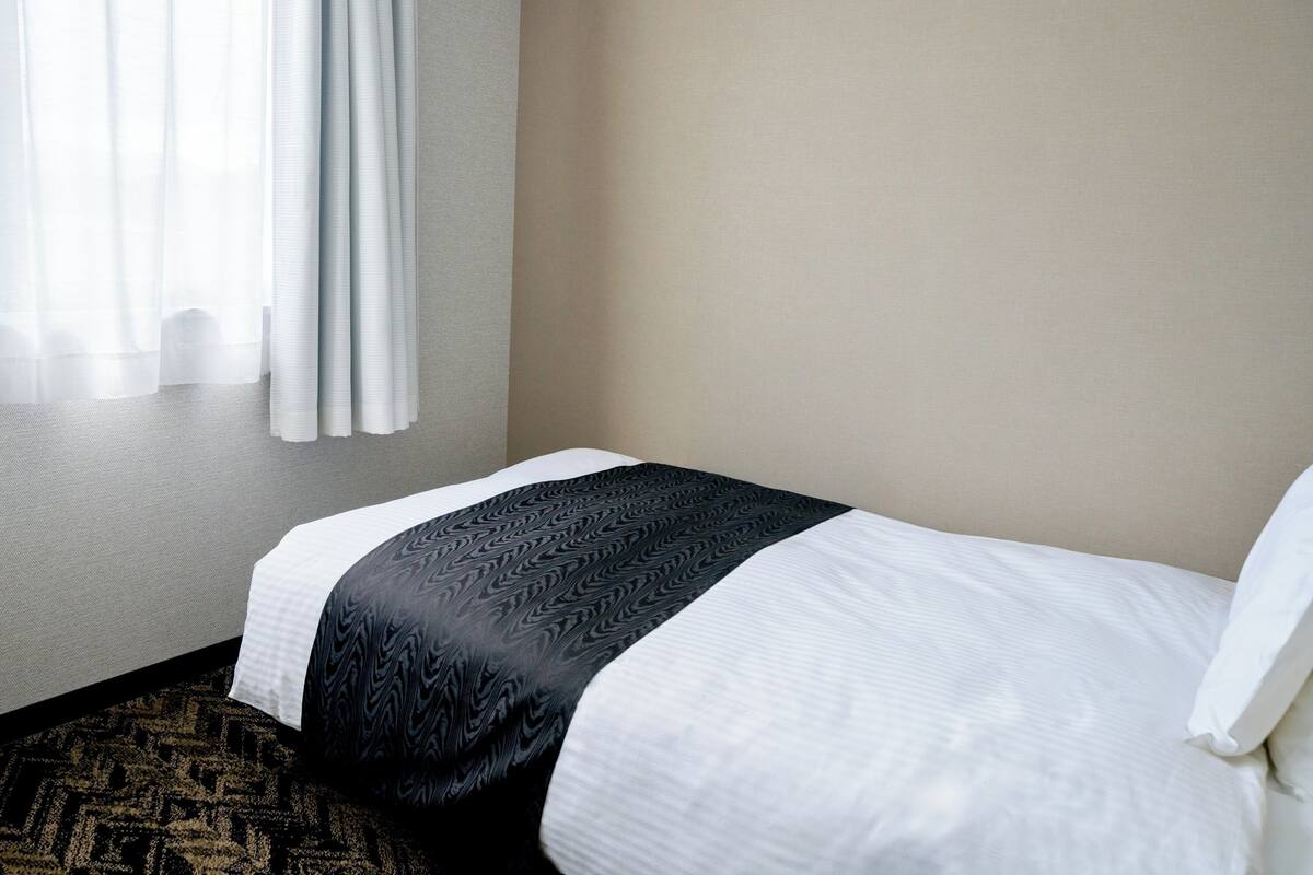 A hotel room with a bed, a window with curtains, and a carpeted floor.