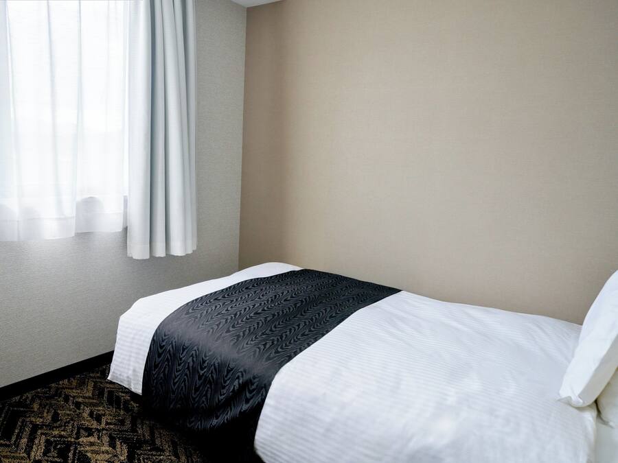 A hotel room with a bed, a window with curtains, and a carpeted floor.