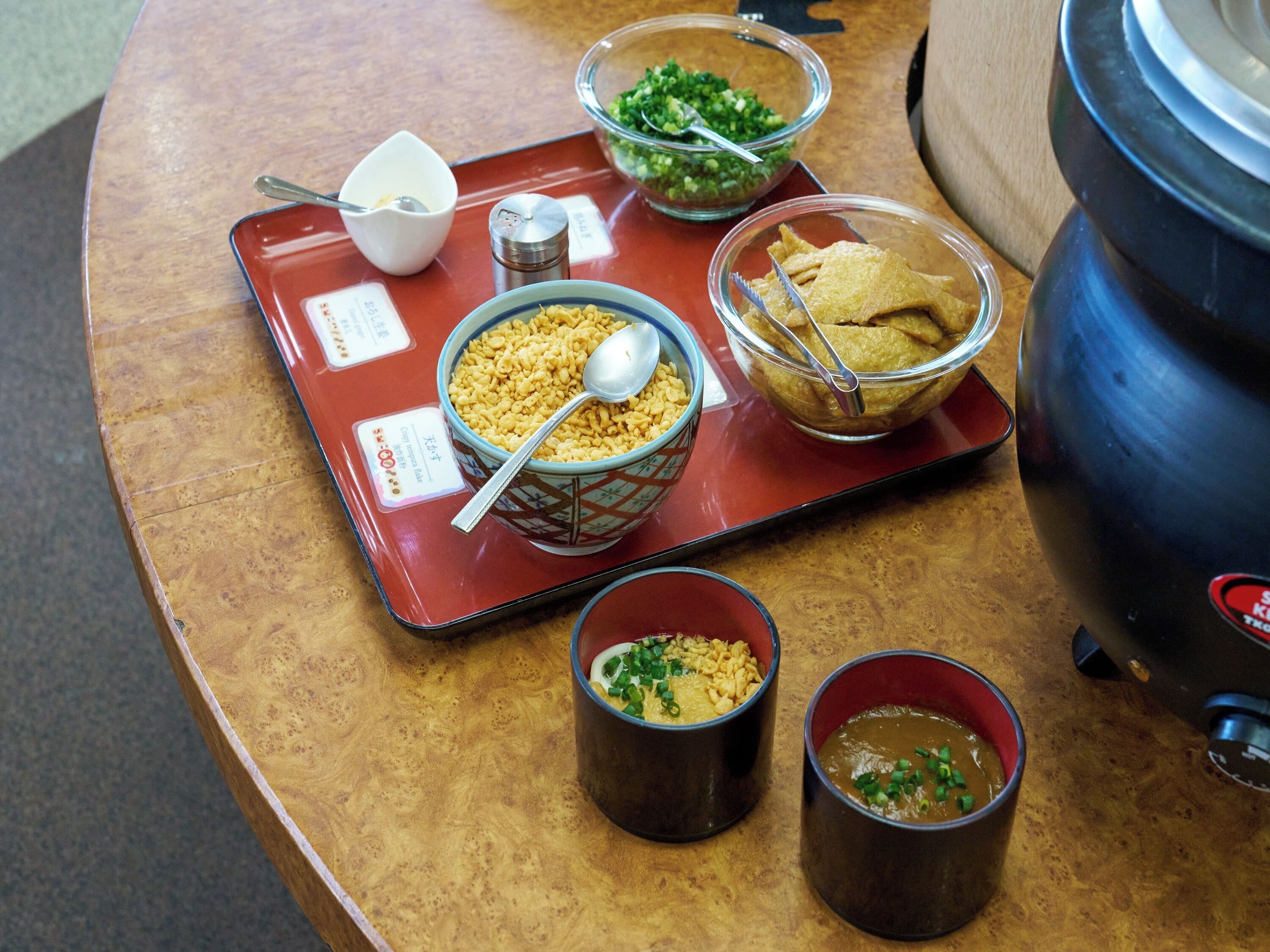 Una mesa con varios platos, incluyendo un tazón de fideos, un tazón de sopa y dos tazas pequeñas, probablemente con salsas o acompañamientos.