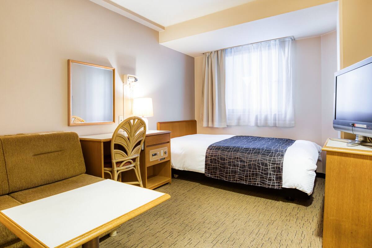 Ein Hotelzimmer mit Bett, Schreibtisch, Stuhl, Fernseher und einem Fenster mit Vorhängen.
