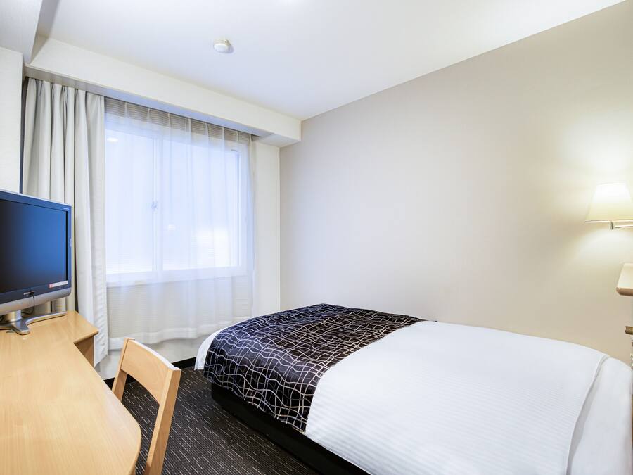 A hotel room with a bed, a desk with a television, a window with curtains, and a wall-mounted lamp.