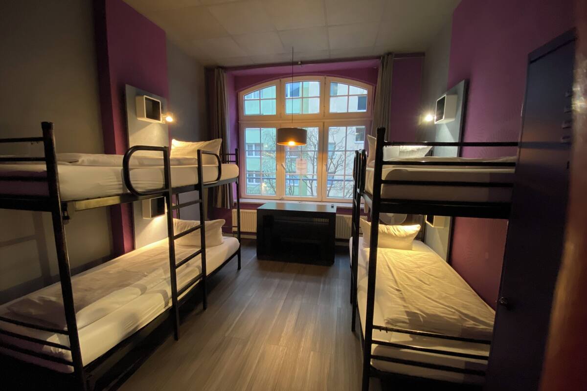 A dormitory room with bunk beds, a window with a view of buildings, and a radiator.