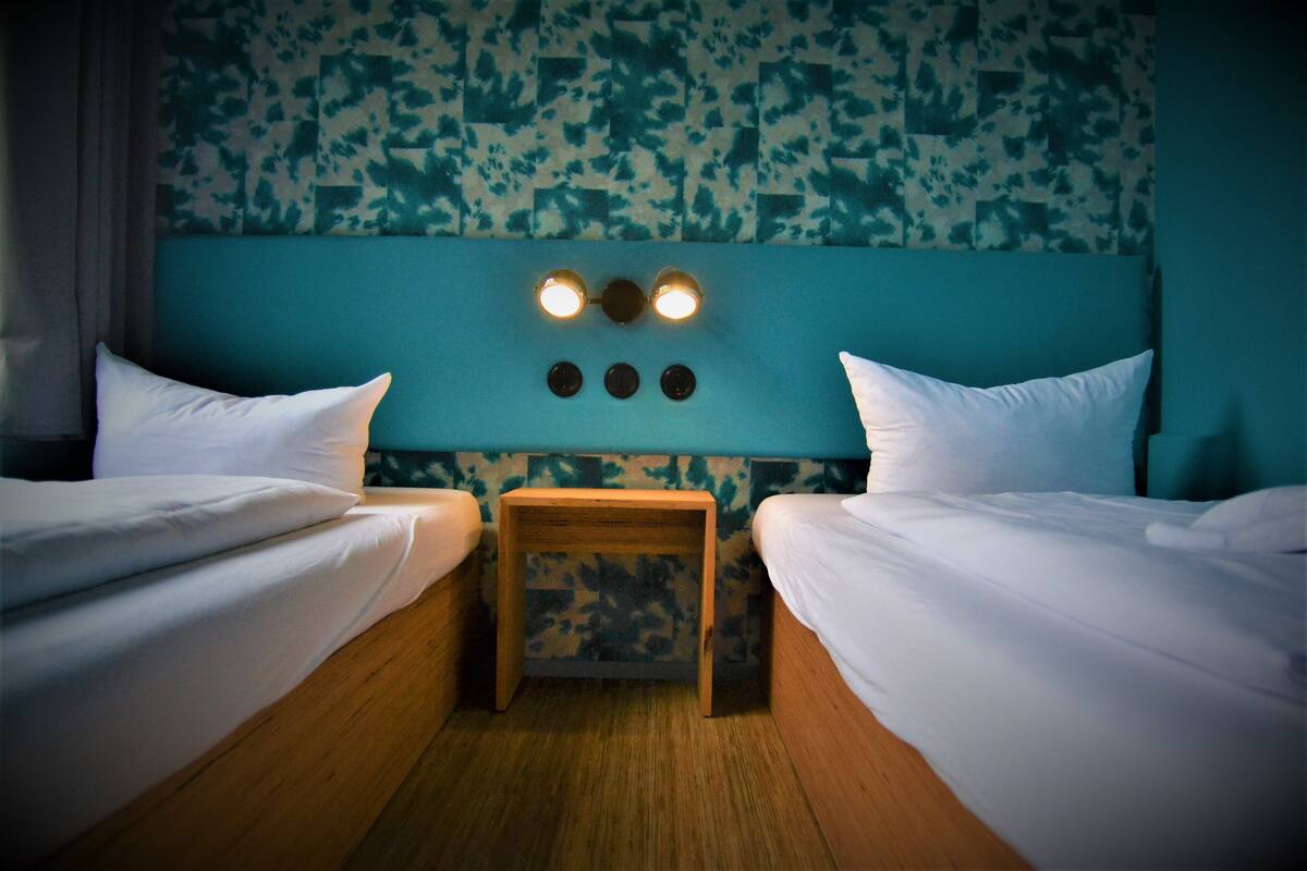 A room with two beds, a small wooden table, and a headboard with wall-mounted lights.