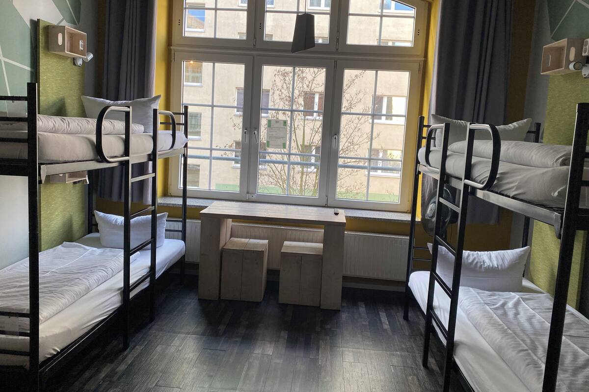 A dormitory room with bunk beds, a desk, and a large window providing a view of buildings and trees.