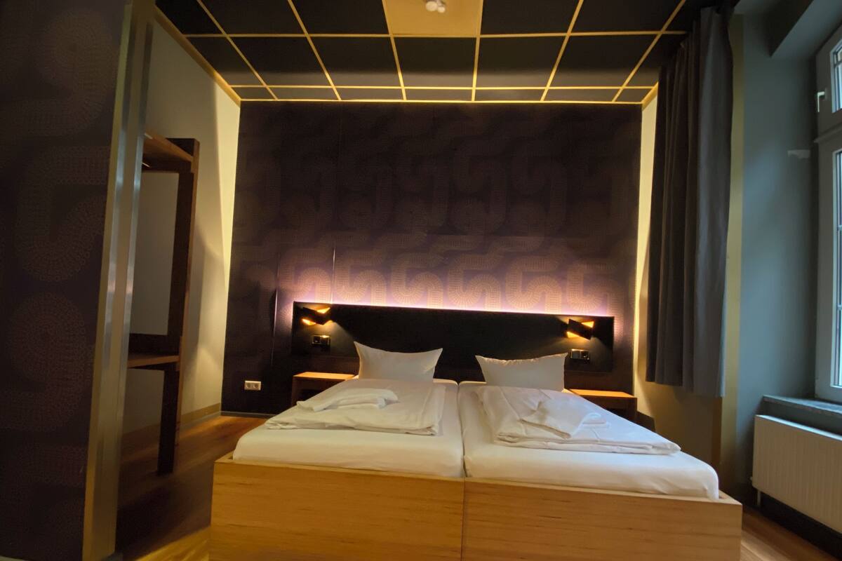 A modern hotel room with a large bed, wooden headboard, and a unique geometric ceiling design.