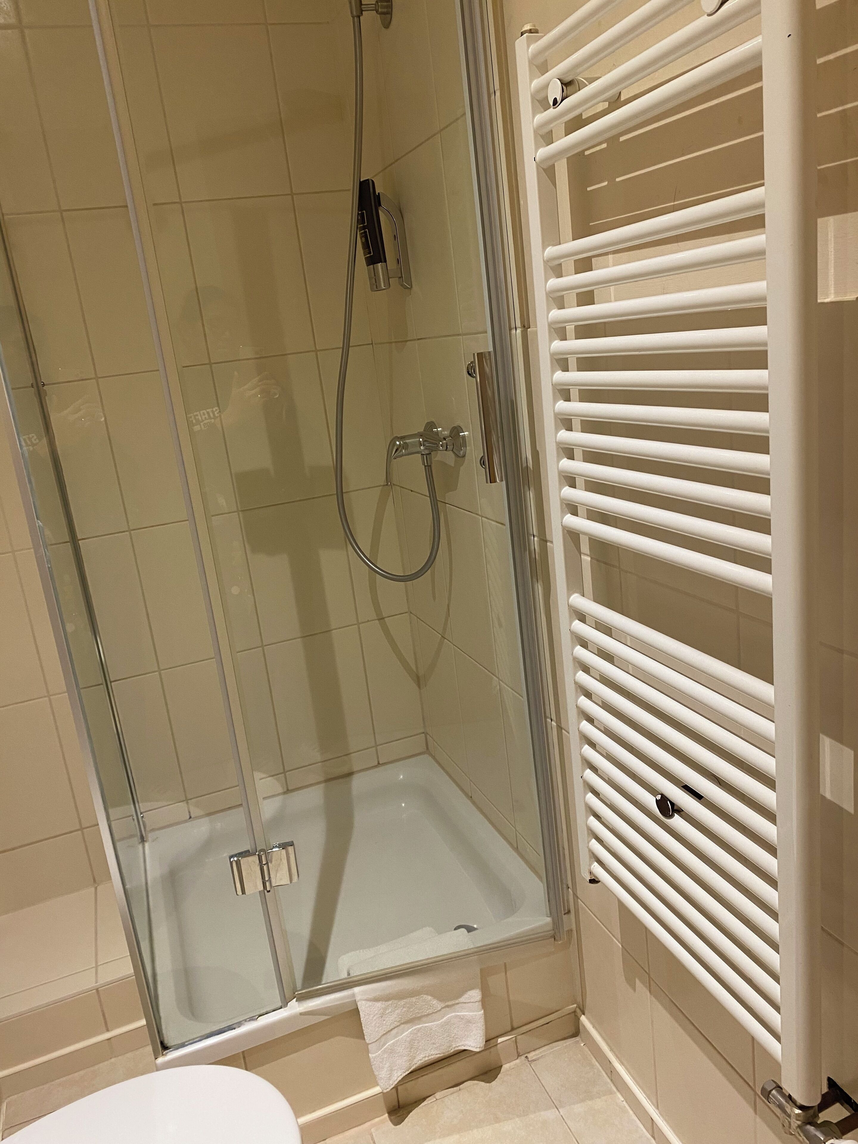 A glass-enclosed shower with a white tiled wall, a white towel on the floor, and a white radiator.