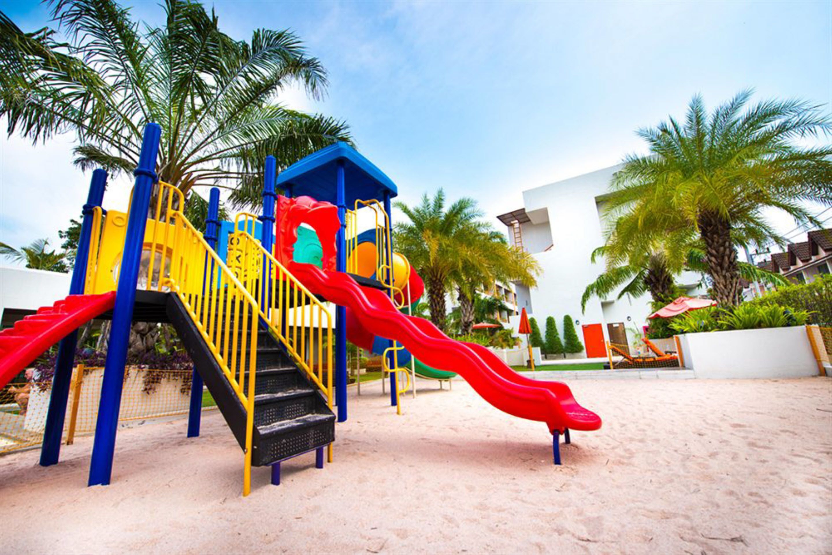 A playground with slides and climbing structures, surrounded by palm trees and a sandy area.