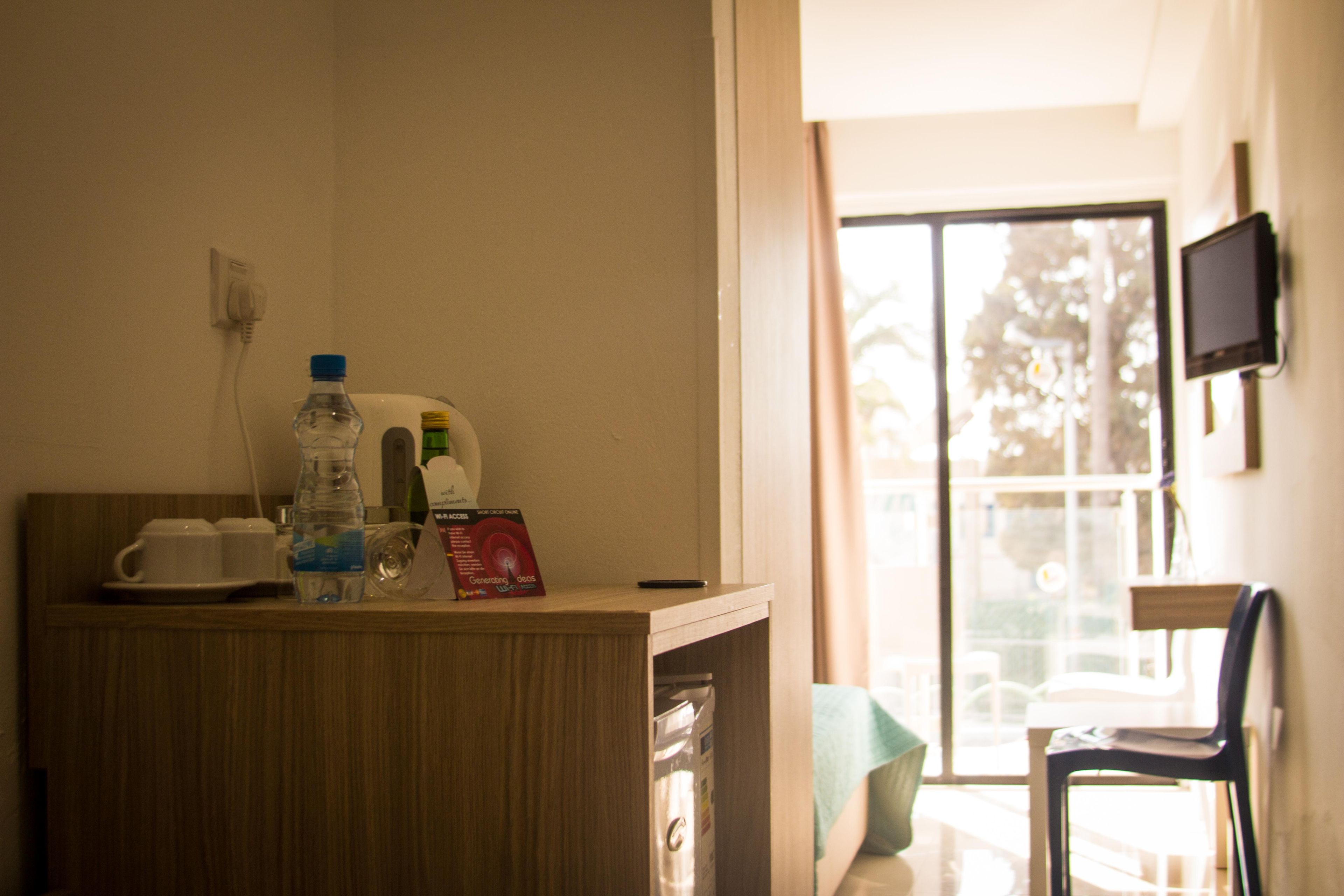 A hotel room with a wooden desk, a water bottle, a cup, a remote control, and a television on the wall.
