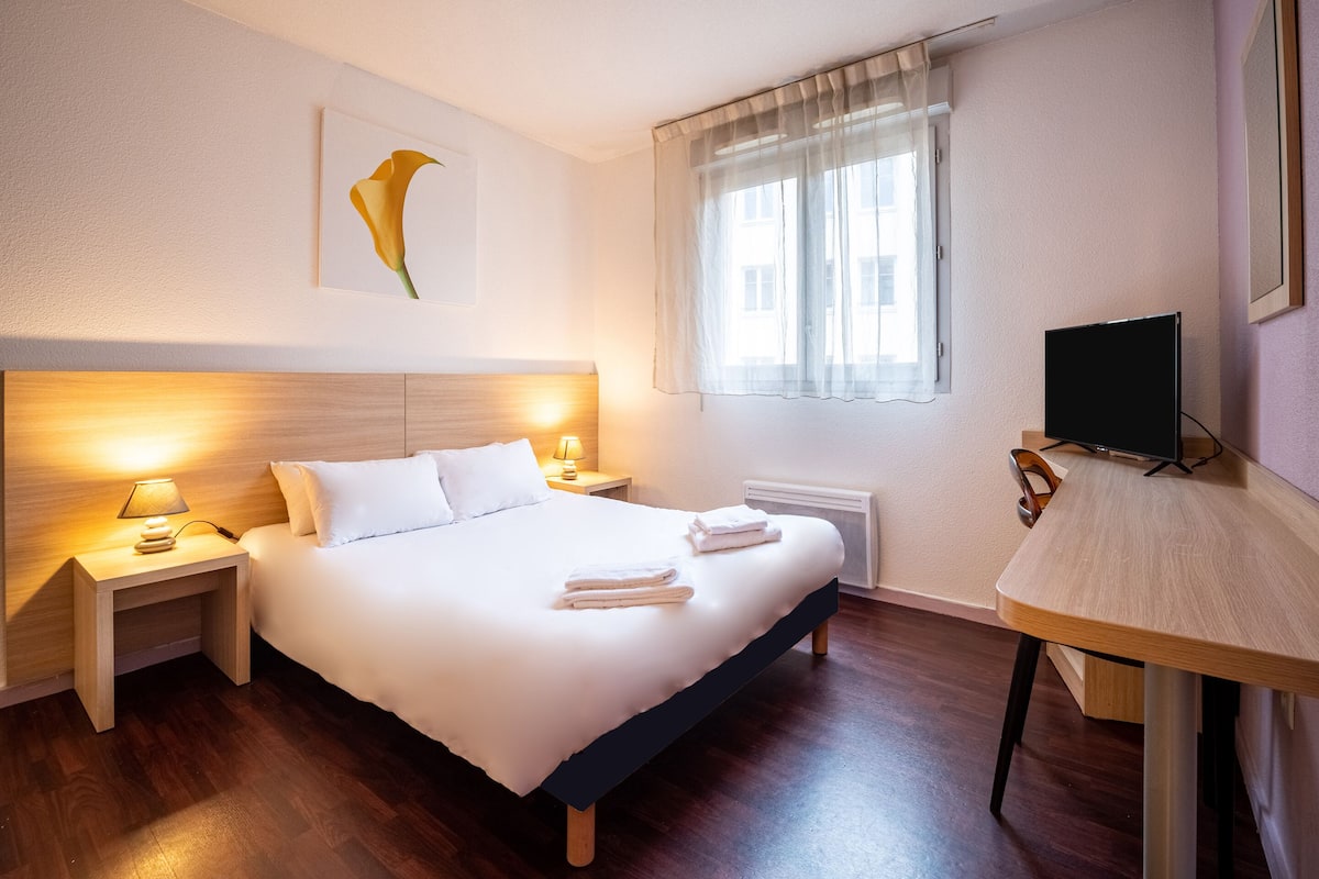 A hotel room with a bed, bedside lamps, a flat-screen TV, a window with sheer curtains, and a wall-mounted artwork.