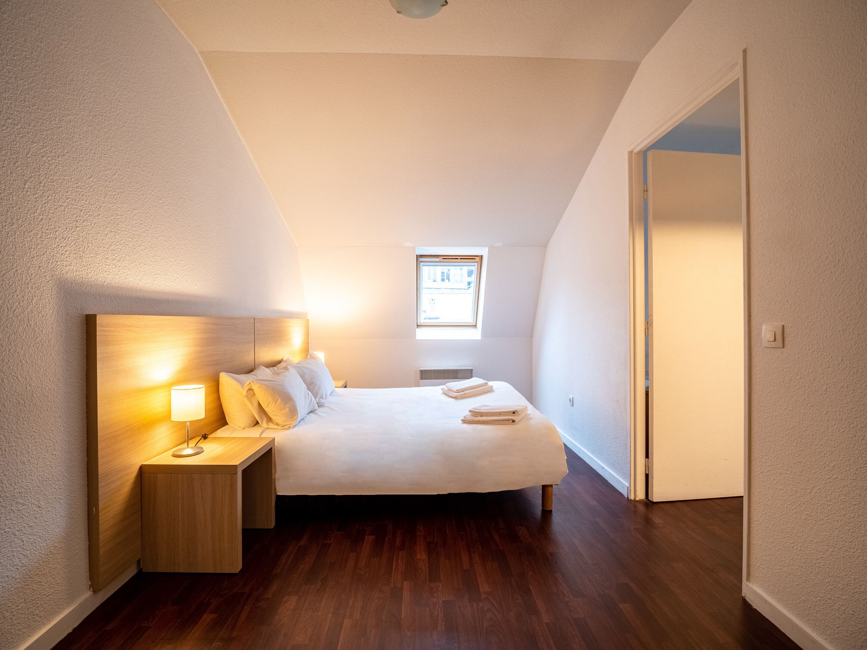 A bedroom with a wooden headboard, a bedside table with a lamp, a neatly made bed, and a window providing natural light.