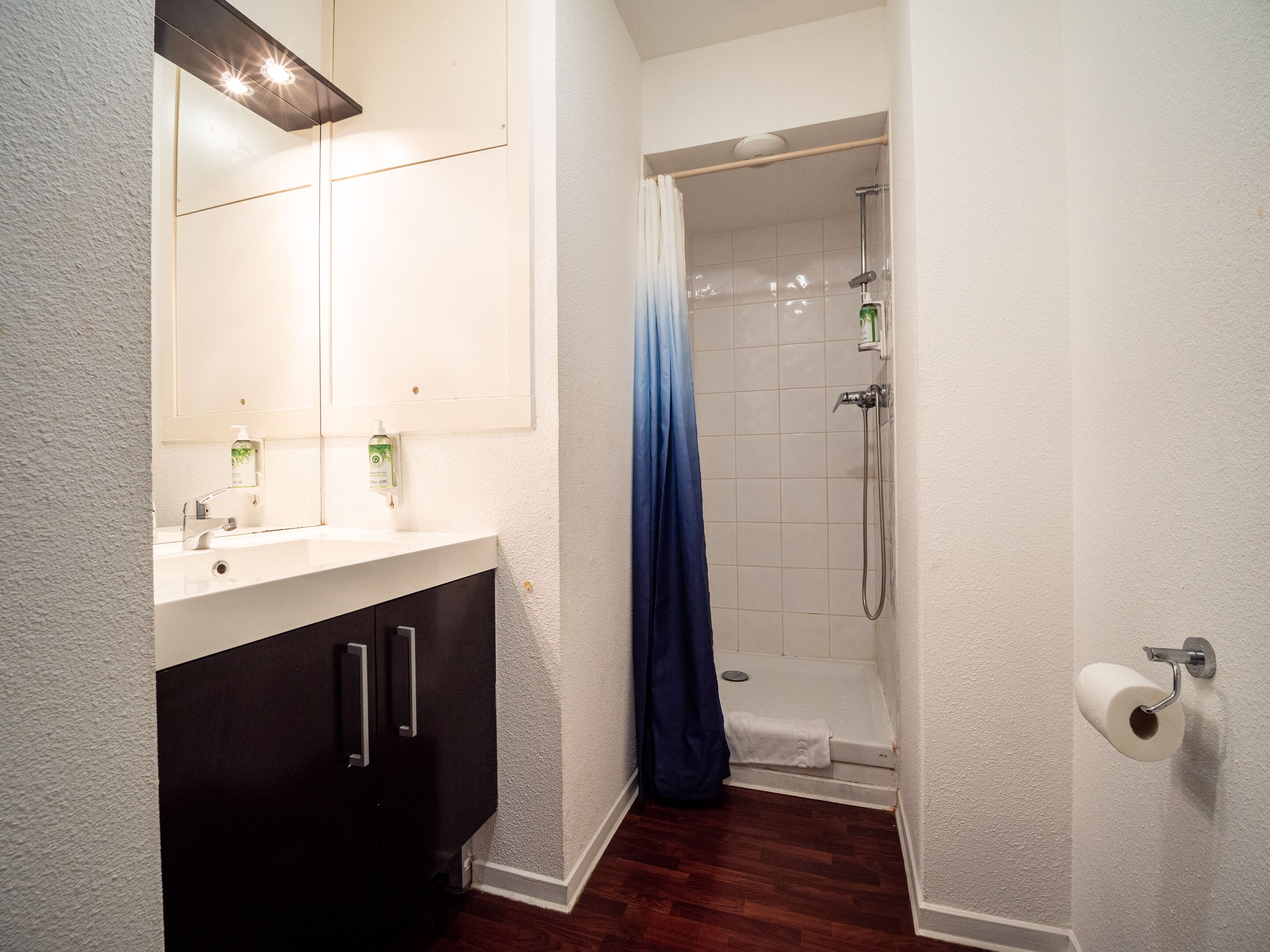 A bathroom with a white sink, dark cabinets, a shower with a blue curtain, and a toilet paper holder.