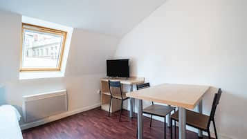 A small, modern room with a sloped ceiling, a wooden desk with a computer, two chairs, a radiator, and a window with a view of buildings.