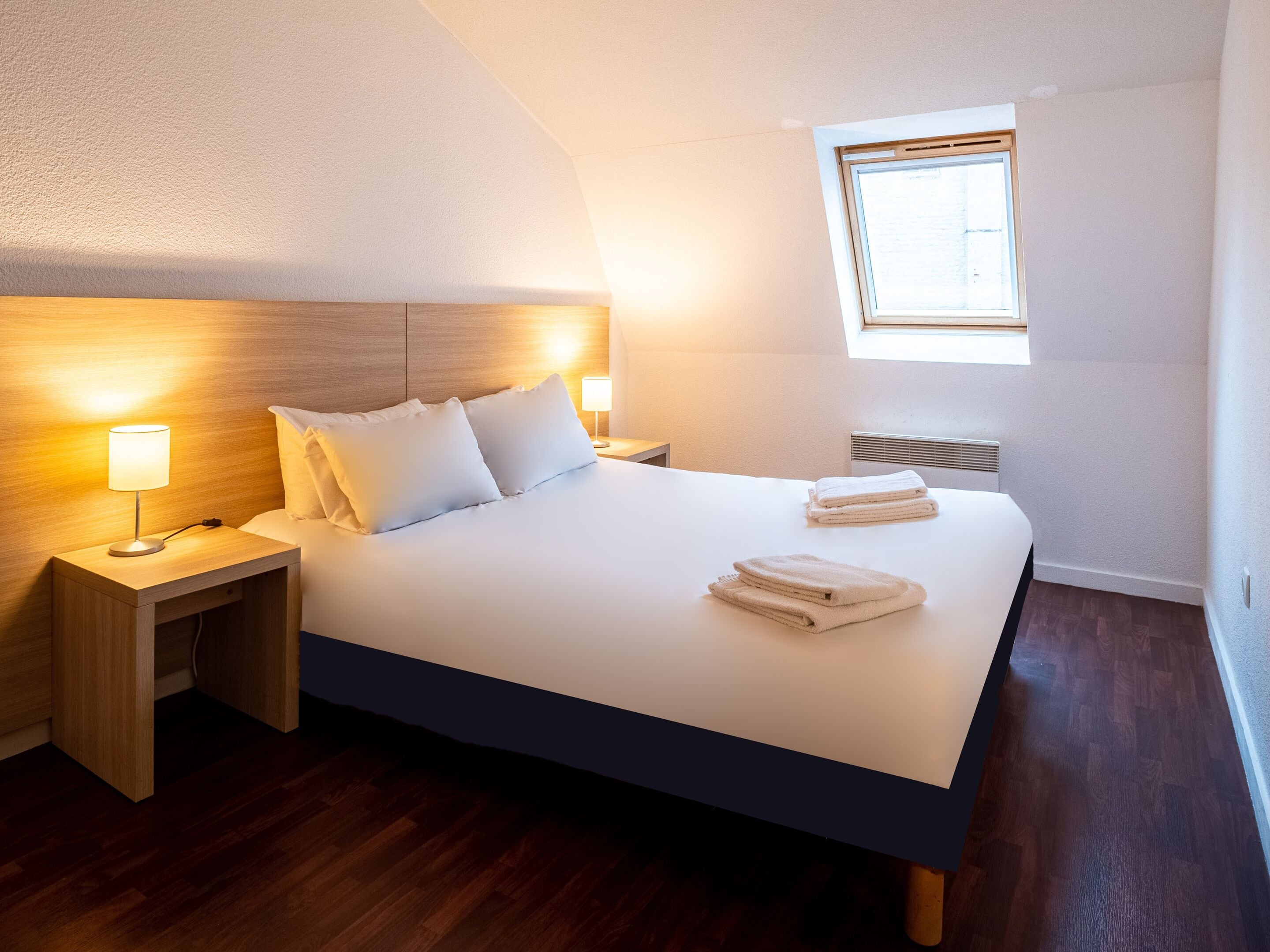 A neatly made bed with white linens and pillows, a bedside table with a lamp, and a window providing natural light.