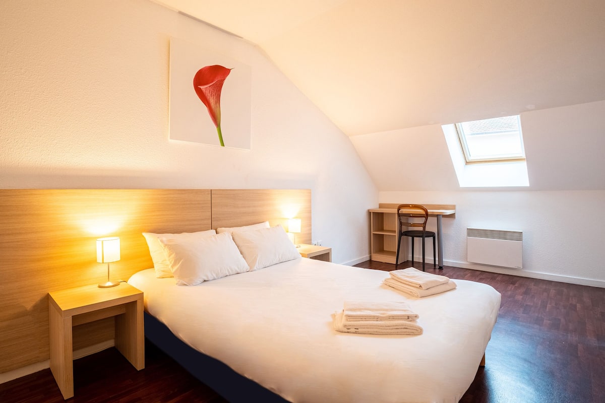 A bedroom with a large bed, a nightstand with a lamp, a desk with a chair, and a skylight.