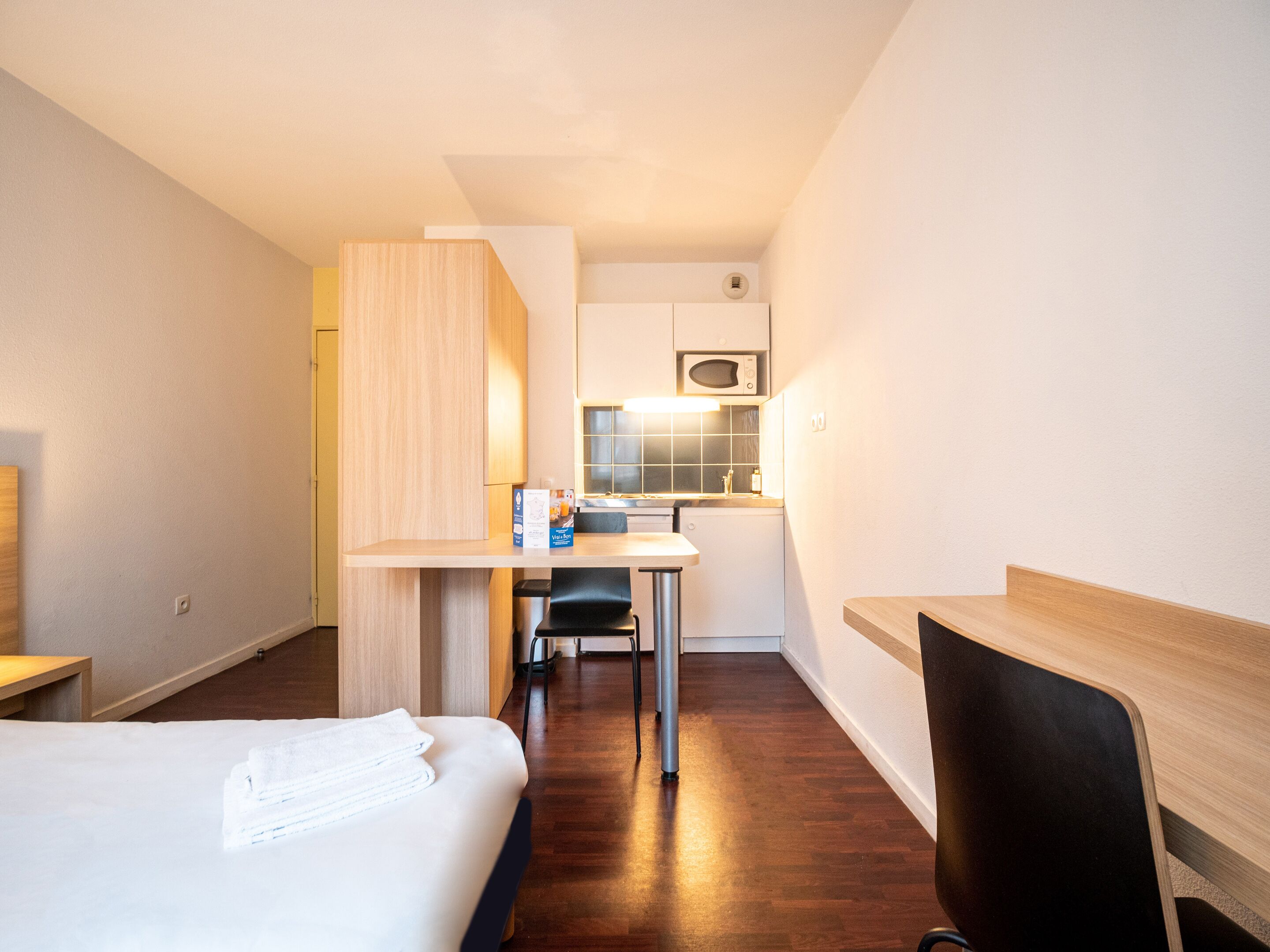 A compact hotel room with a bed, a desk, a chair, a small kitchenette, and a wooden wardrobe.