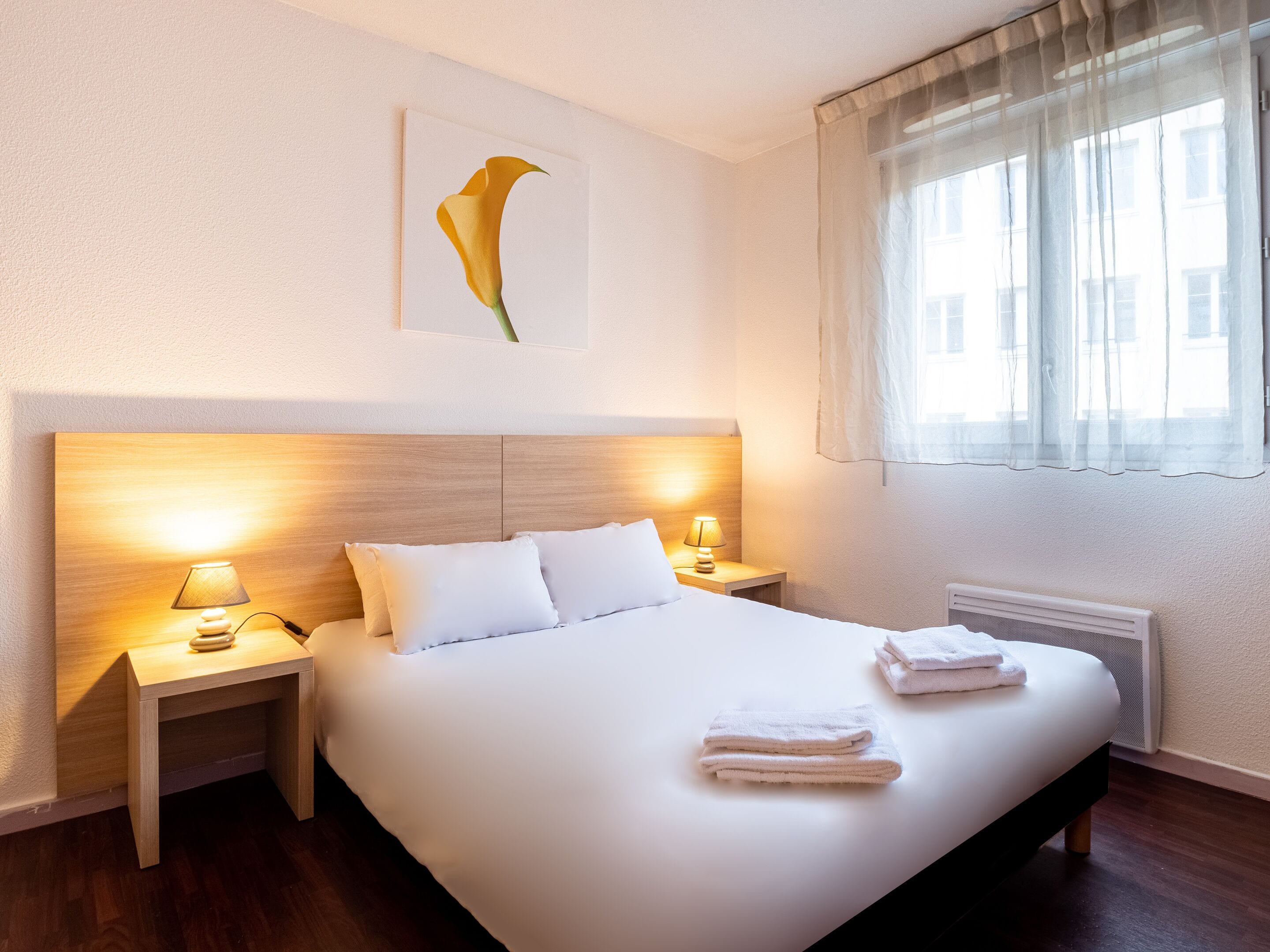 A hotel room with a large bed, two bedside tables with lamps, a radiator, and a window with sheer curtains.