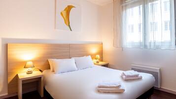A hotel room with a large bed, two bedside tables with lamps, a radiator, and a window with sheer curtains.