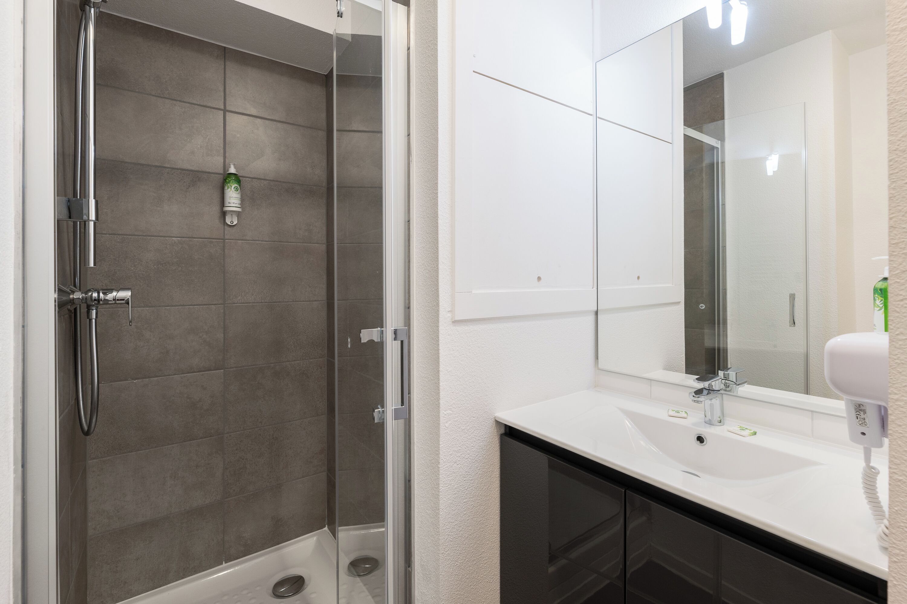 Un baño moderno con ducha envuelta en vidrio, un lavamanos blanco con espejo y un secador de pelo en la encimera.