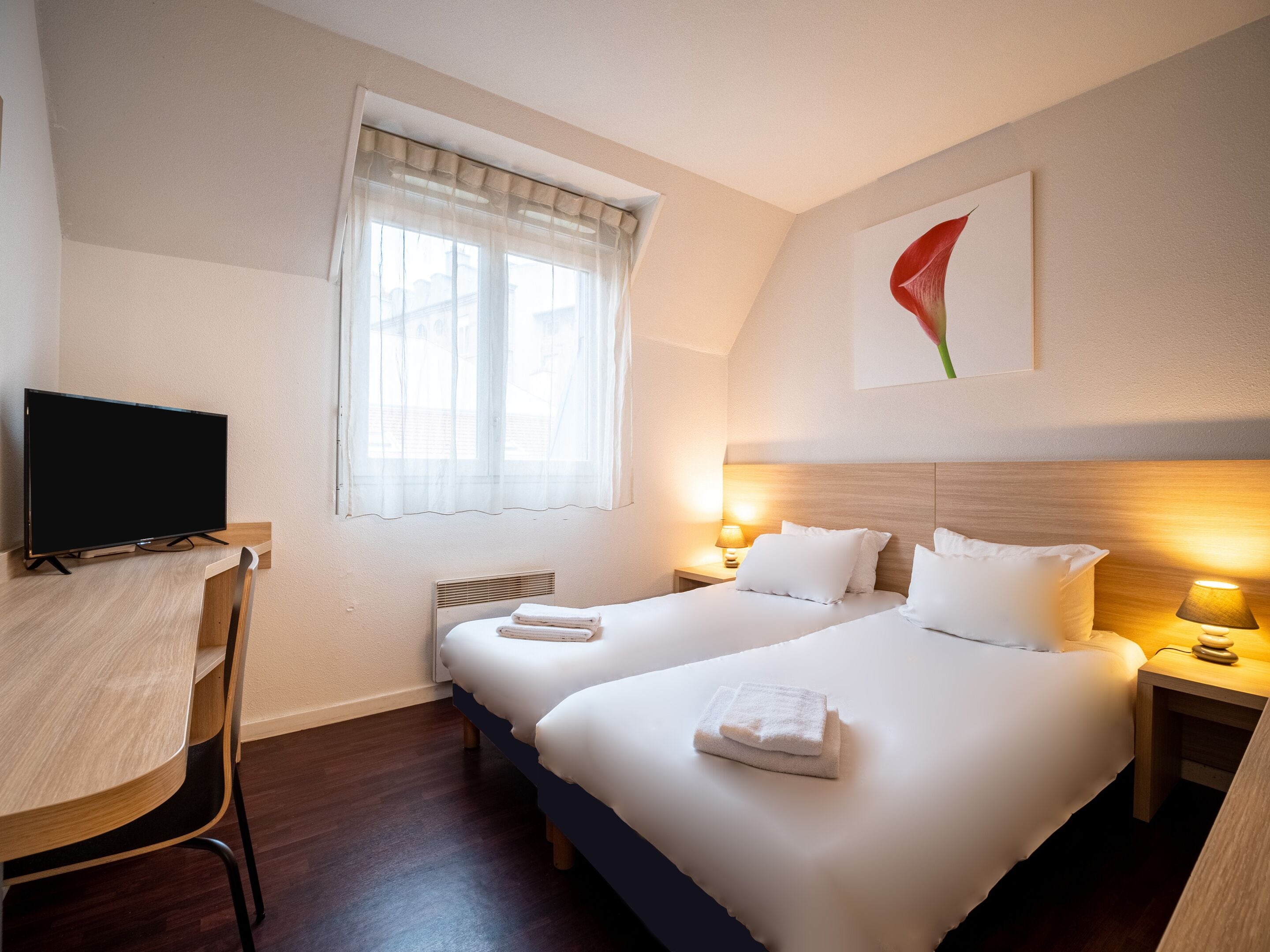 A hotel room with two beds, a desk, a TV, and a window with sheer curtains.