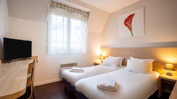 A hotel room with two beds, a desk, a TV, and a window with sheer curtains.