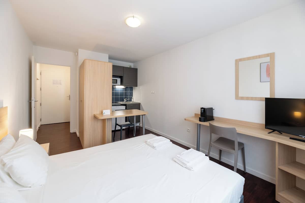 A hotel room with a bed, a desk with a TV, a kitchenette, and a bathroom.