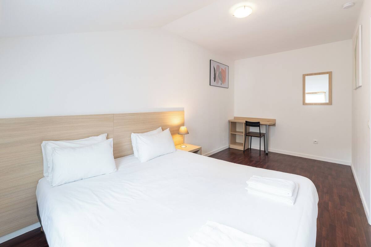 A neatly made bed with white linens and pillows, a bedside lamp, a small wooden desk with a chair, and a framed picture on the wall.