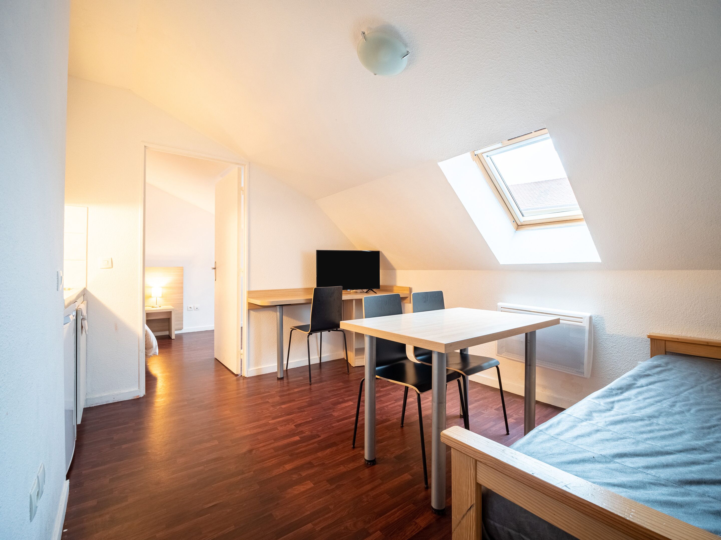 A small, well-lit room with a wooden floor, a bed, a desk with a chair, and a TV.