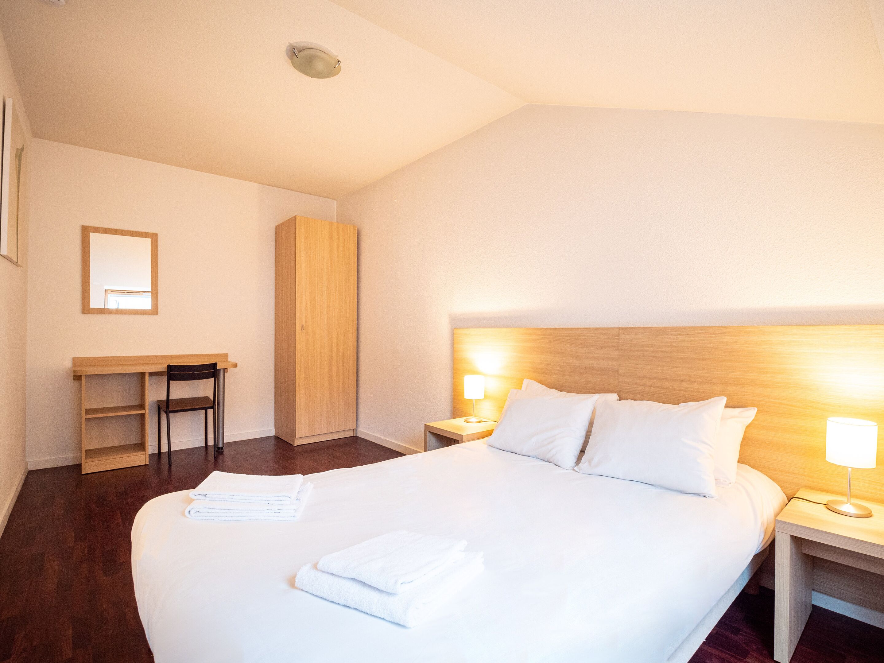 A hotel room with a large bed, a desk, a chair, and a wardrobe.