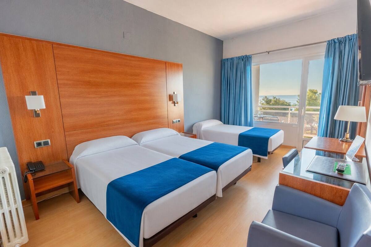 A hotel room with two beds, a desk, a chair, a radiator, and a window with curtains.