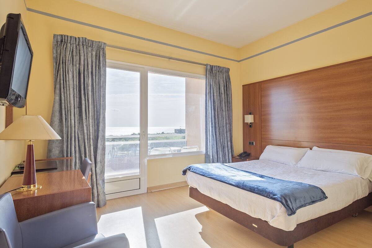 A hotel room with a bed, a desk, a chair, a TV, and a view of the sea.