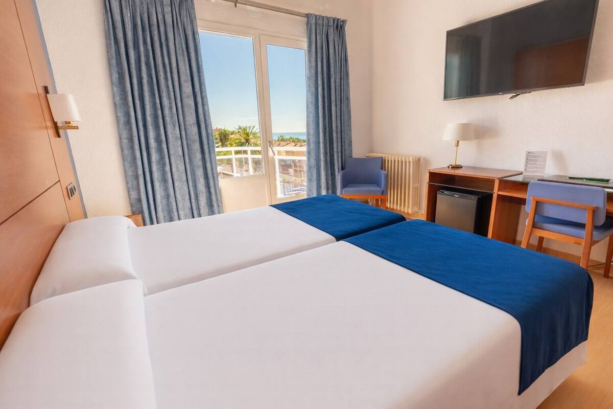 A hotel room with two beds, a desk, a chair, a TV, and a view of the sea.