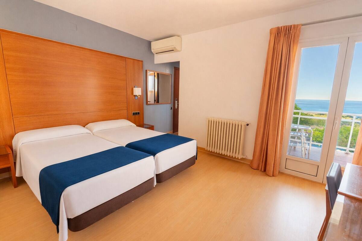 A hotel room with two beds, a wooden headboard, a radiator, a sliding glass door with a view, and a small table.