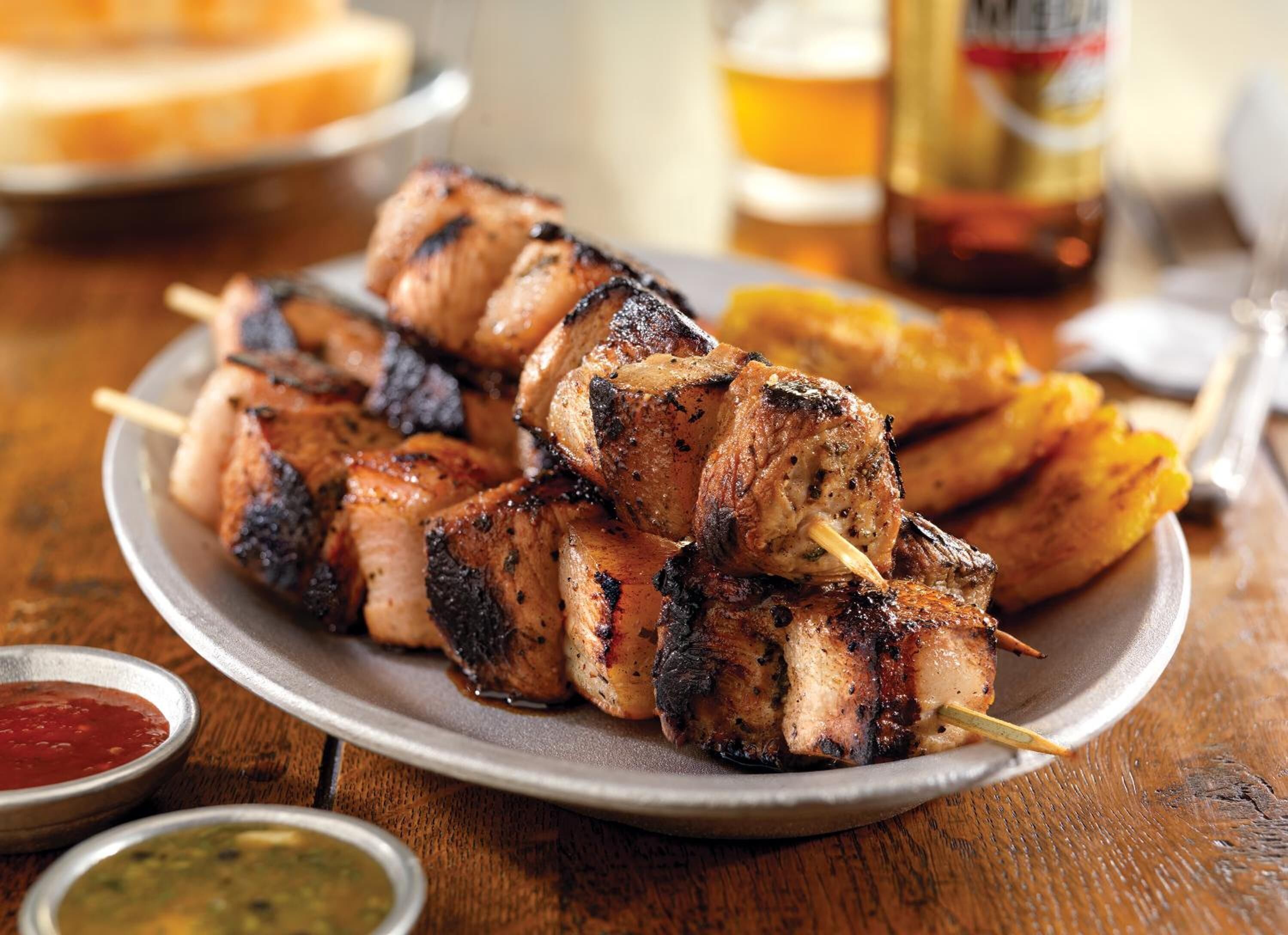 A plate of grilled meat skewers with a side of fried plantains and two dipping sauces.