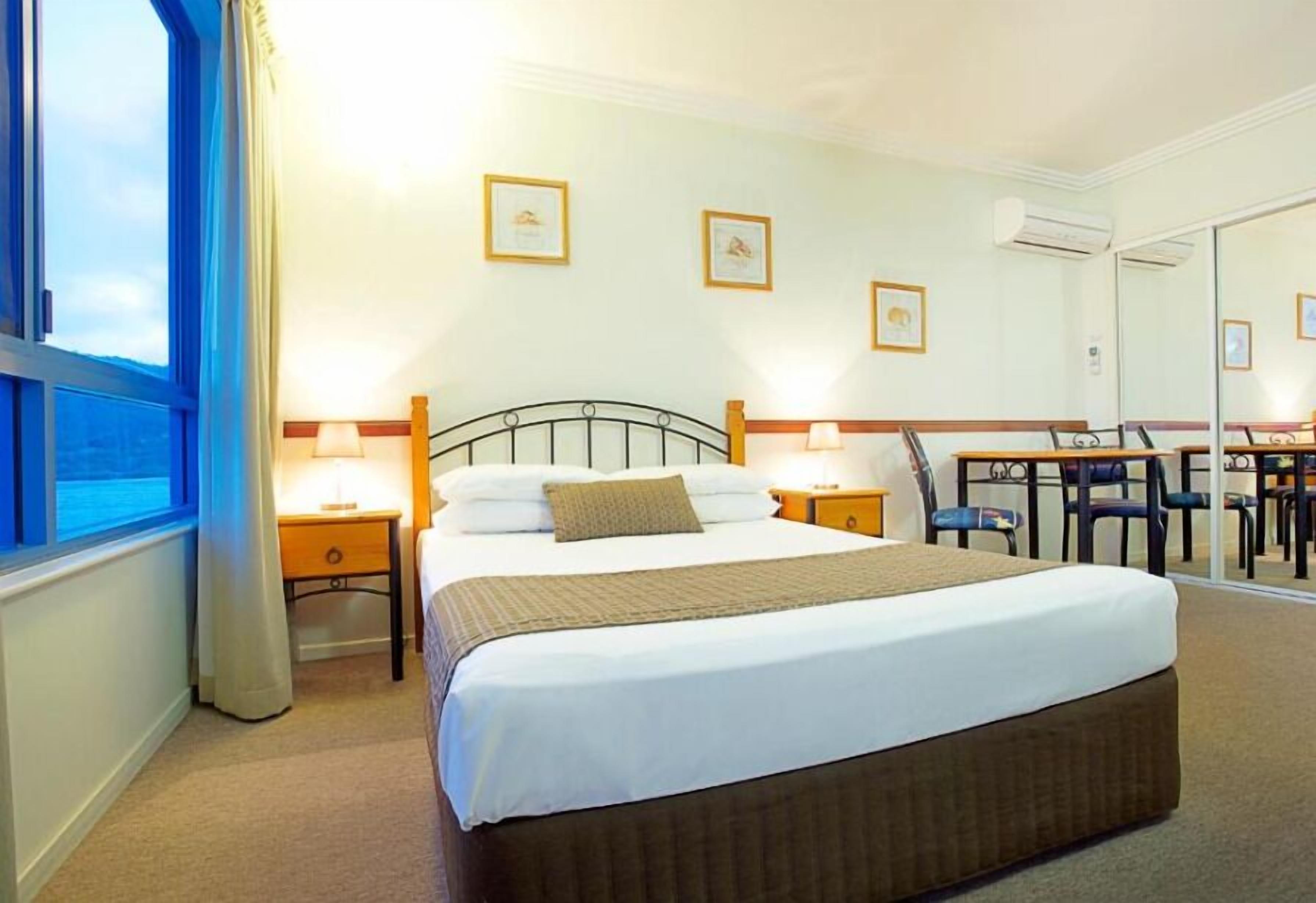 A hotel room with a large bed, bedside tables with lamps, a dining area with chairs, and framed pictures on the wall.