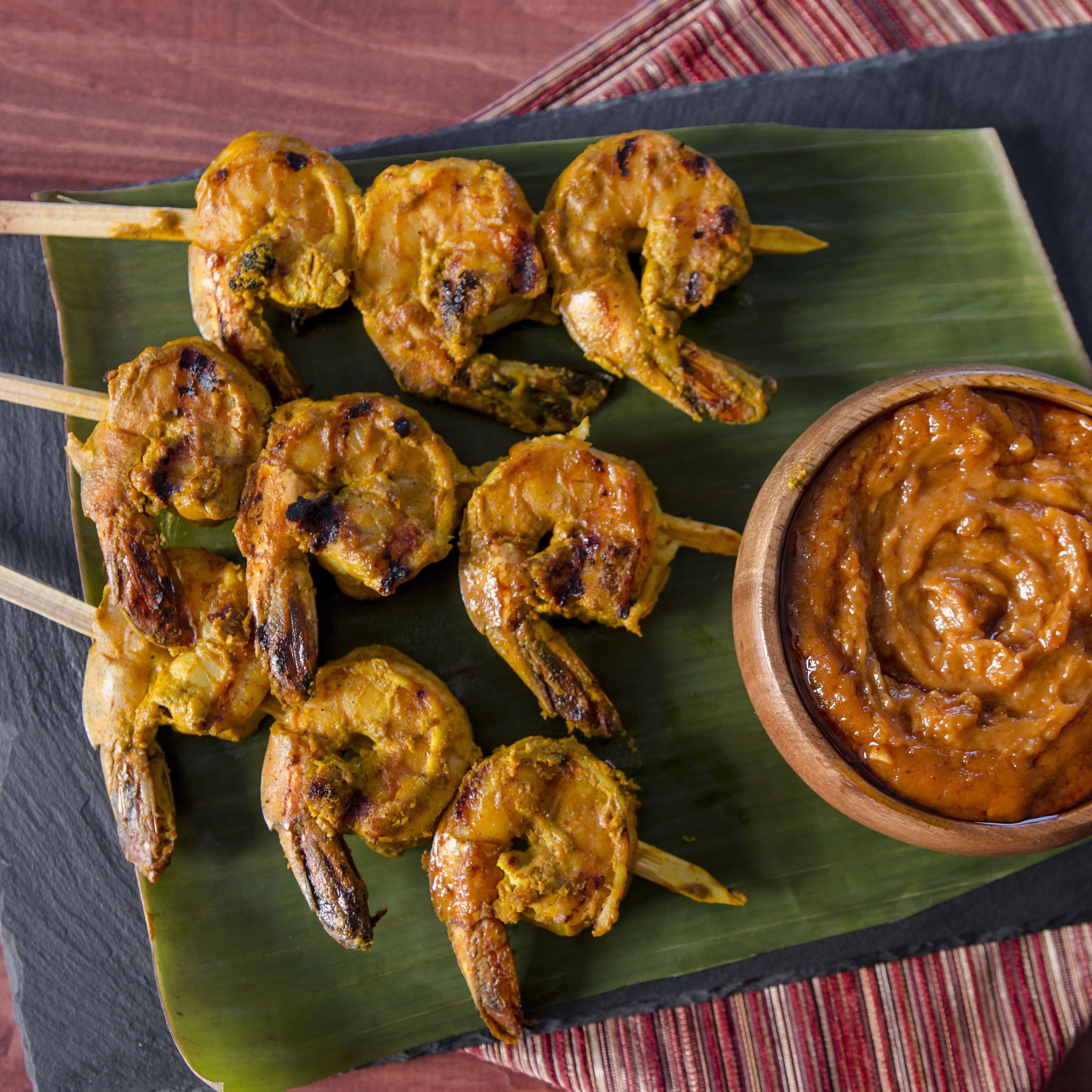 Skewered grilled shrimp on a banana leaf with a bowl of sauce.