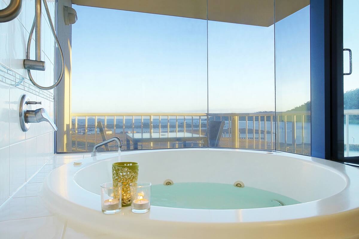 A modern bathroom with a large bathtub, a glass shower enclosure, and a view of a bridge and cityscape.