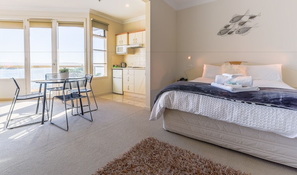 A bedroom with a bed, bedside table, and a view of the kitchen and balcony.