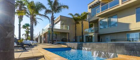 A modern hotel with a swimming pool, palm trees, and a clear blue sky.