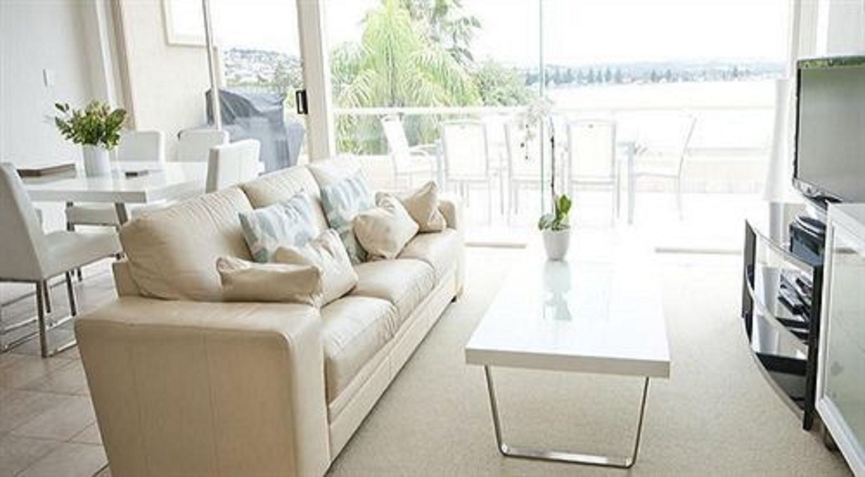 A modern living room with a sofa, a coffee table, and a TV. There is a balcony with outdoor furniture and a view of the outdoors.