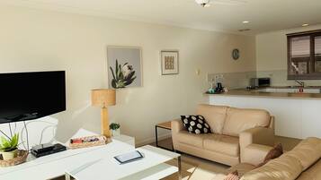 A modern living room with a flat-screen TV, a white coffee table, a beige sofa, and a small kitchen area with a microwave.