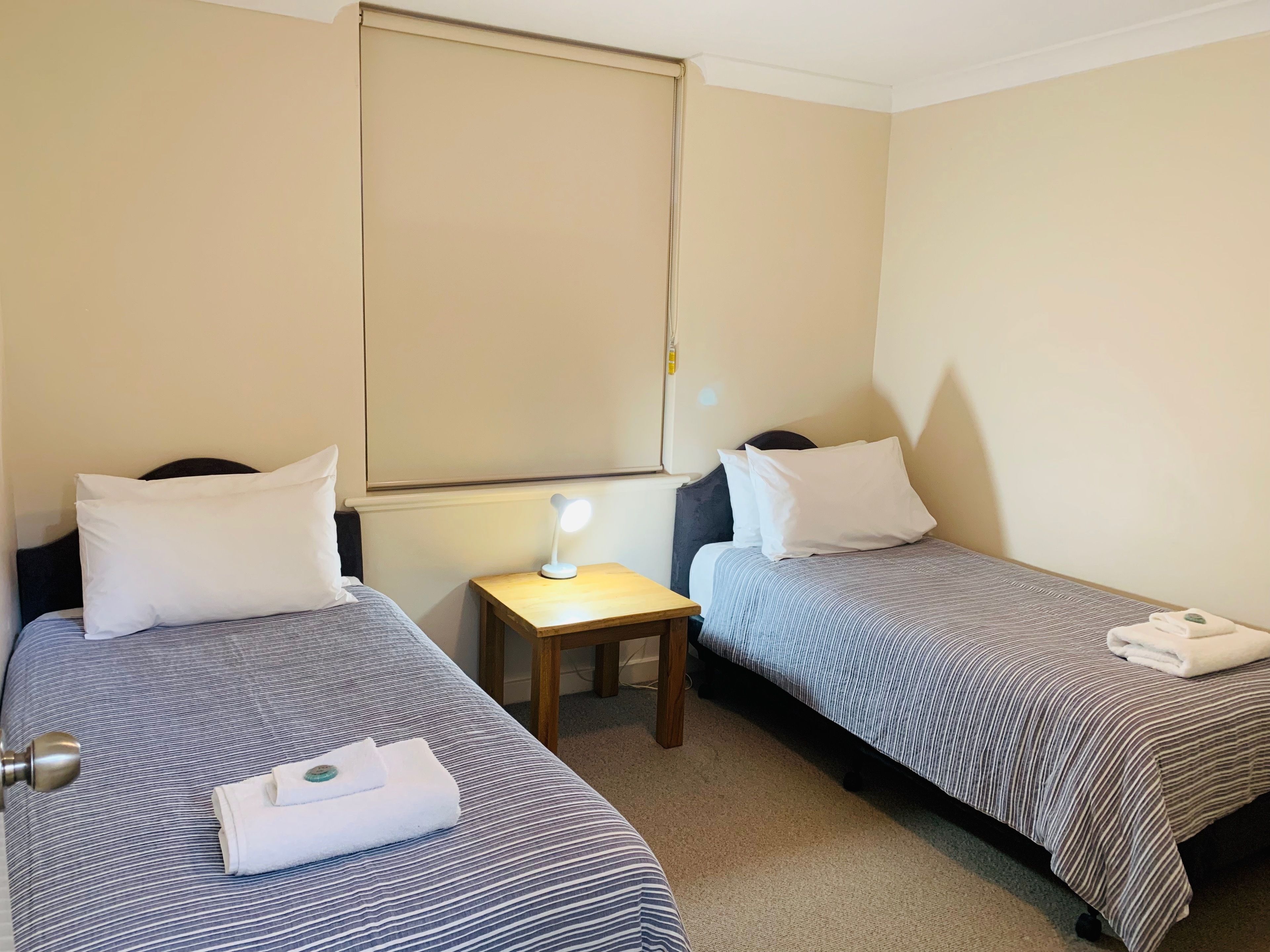 A hotel room with two single beds, each with a striped bedspread and white pillows. There is a small wooden nightstand with a lamp between the beds. A folded towel is on the bed on the right.