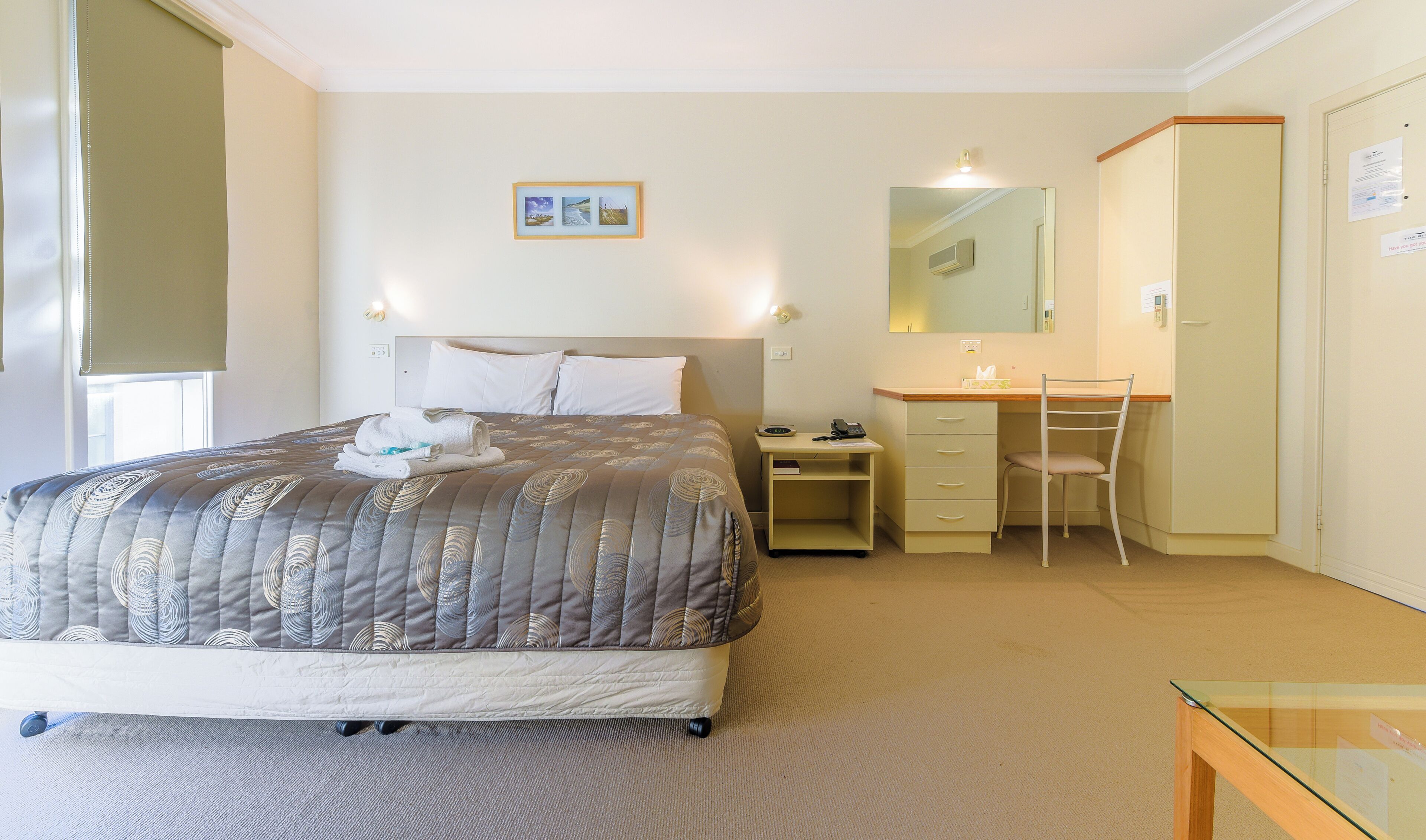 A hotel room with a bed, a desk with a chair, a mirror, and a small table.