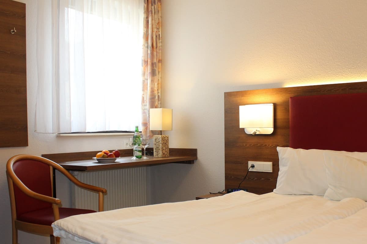 A hotel room with a bed, a desk with a lamp, a chair, and a window with curtains.