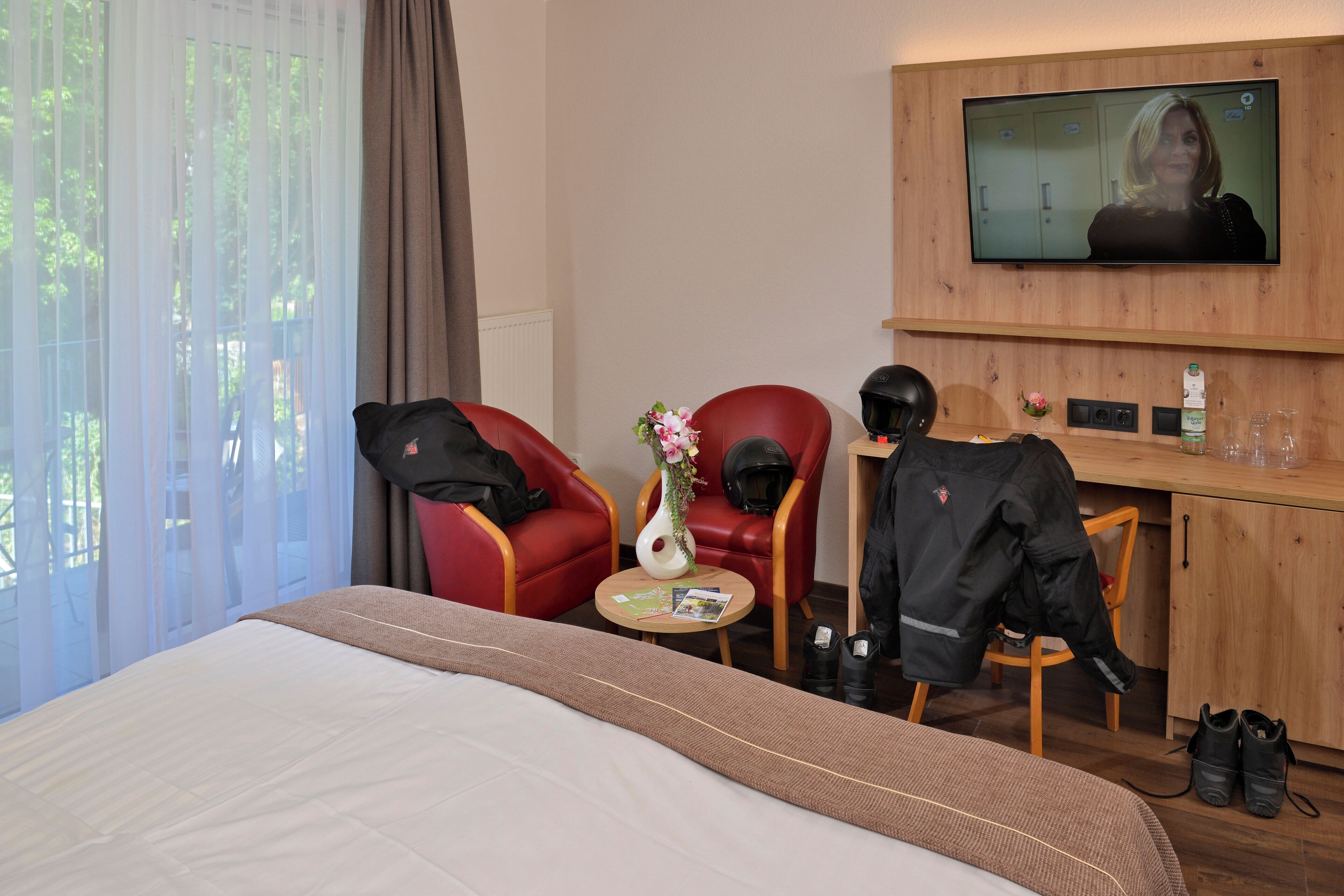 Hotelkamer met een bed, een bureau met een televisie, een rode stoel, een witte vaas met bloemen en een houten kast.